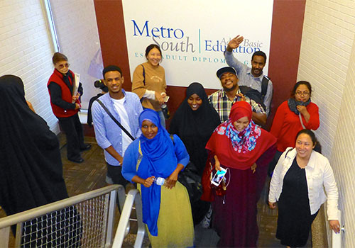 a group of students, some wearing the hijab or khimar stand in a stairwell with the Metro South Adult Basic Education logo on the wall behind them